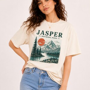 Pode incluir: T-shirt creme com um design de paisagem e o texto "JASPER - NATIONAL PARK". O gr&aacute;fico inclui um rio, montanhas, &aacute;rvores e um sol poente. A camisa &eacute; combinada com jeans azul claro.