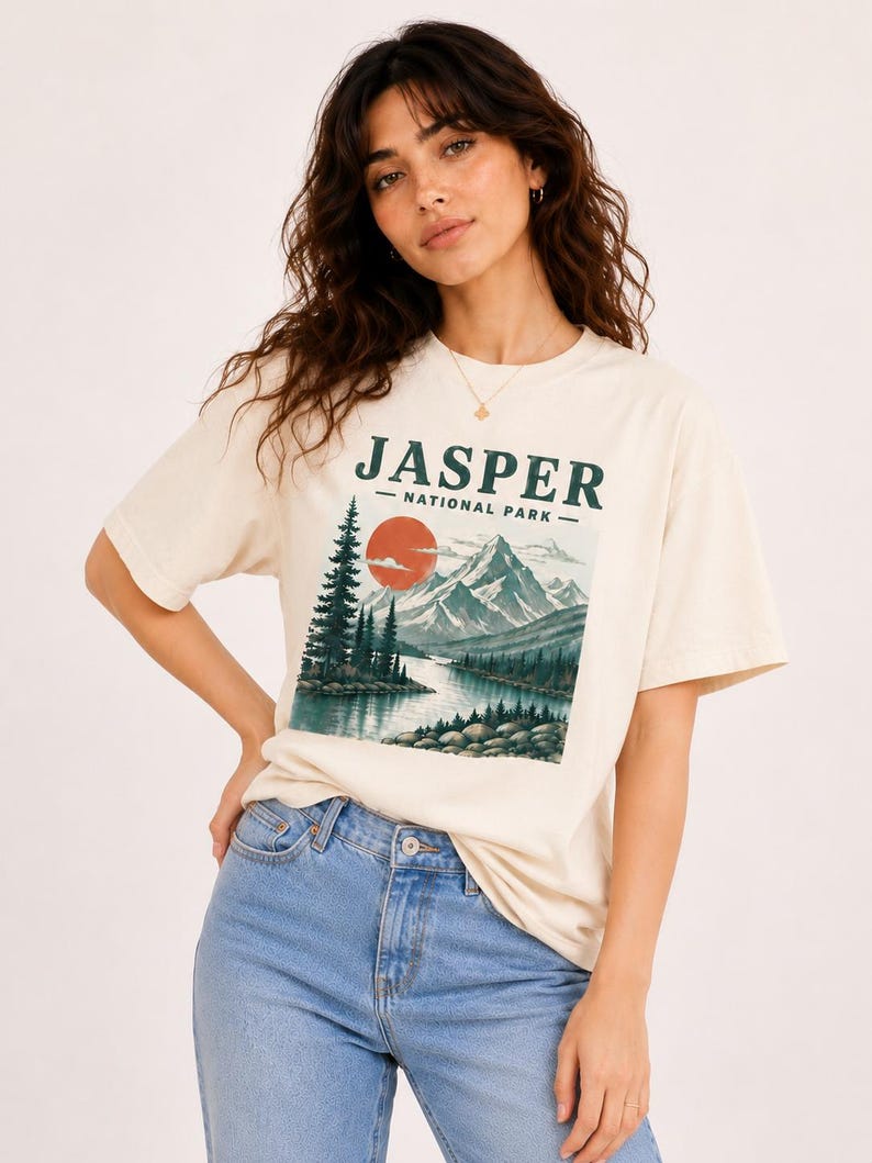 Pode incluir: T-shirt creme com um design de paisagem e o texto "JASPER - NATIONAL PARK". O gr&aacute;fico inclui um rio, montanhas, &aacute;rvores e um sol poente. A camisa &eacute; combinada com jeans azul claro.