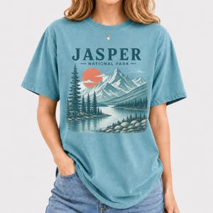 Pode incluir: Uma camiseta azul-petr&oacute;leo com uma ilustra&ccedil;&atilde;o c&ecirc;nica do Parque Nacional Jasper. O design inclui montanhas, um lago, &aacute;rvores e um sol poente. O texto "JASPER NATIONAL PARK" est&aacute; impresso acima da paisagem.