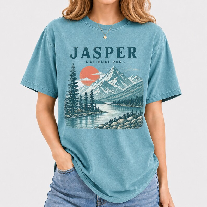 Pode incluir: Uma camiseta azul-petr&oacute;leo com uma ilustra&ccedil;&atilde;o c&ecirc;nica do Parque Nacional Jasper. O design inclui montanhas, um lago, &aacute;rvores e um sol poente. O texto "JASPER NATIONAL PARK" est&aacute; impresso acima da paisagem.