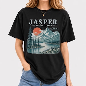 Pode incluir: Camiseta preta com um design de paisagem. O gr&aacute;fico inclui o texto "JASPER - NATIONAL PARK" acima de uma cordilheira, um lago e &aacute;rvores, com um sol poente. A camiseta tem um corte folgado.