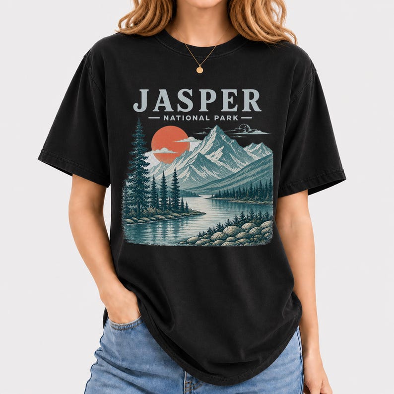 Pode incluir: Camiseta preta com um design de paisagem. O gr&aacute;fico inclui o texto "JASPER - NATIONAL PARK" acima de uma cordilheira, um lago e &aacute;rvores, com um sol poente. A camiseta tem um corte folgado.