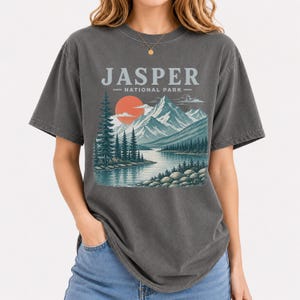 Pode incluir: T-shirt cinzenta escura com uma ilustra&ccedil;&atilde;o do Parque Nacional de Jasper. O design inclui montanhas, um lago, &aacute;rvores e um sol poente. O texto "JASPER - NATIONAL PARK -" est&aacute; impresso acima da paisagem.