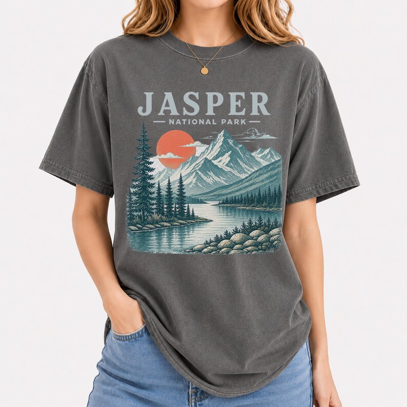Pode incluir: T-shirt cinzenta escura com uma ilustra&ccedil;&atilde;o do Parque Nacional de Jasper. O design inclui montanhas, um lago, &aacute;rvores e um sol poente. O texto "JASPER - NATIONAL PARK -" est&aacute; impresso acima da paisagem.