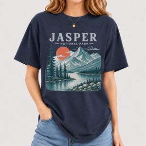 Pode incluir: Camiseta azul marinho com uma ilustra&ccedil;&atilde;o do Parque Nacional Jasper. O design inclui uma cordilheira, um lago e um p&ocirc;r do sol. O texto "JASPER - NATIONAL PARK" est&aacute; impresso acima da paisagem.