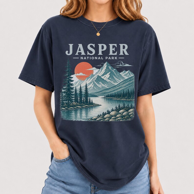Pode incluir: Camiseta azul marinho com uma ilustra&ccedil;&atilde;o do Parque Nacional Jasper. O design inclui uma cordilheira, um lago e um p&ocirc;r do sol. O texto "JASPER - NATIONAL PARK" est&aacute; impresso acima da paisagem.