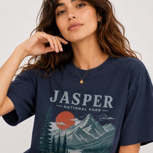 Pode incluir: Camiseta azul marinho com o texto "JASPER NATIONAL PARK" acima de um gr&aacute;fico de montanhas, lago e p&ocirc;r do sol. A camiseta tem mangas curtas e gola redonda.