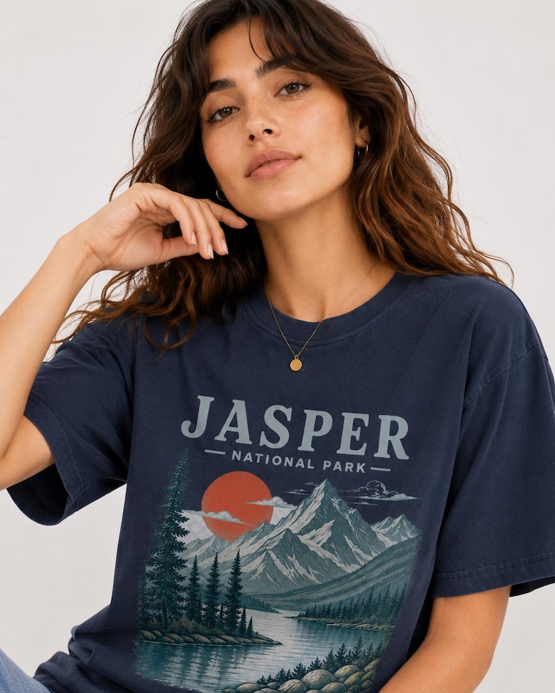 Pode incluir: Camiseta azul marinho com o texto "JASPER NATIONAL PARK" acima de um gr&aacute;fico de montanhas, lago e p&ocirc;r do sol. A camiseta tem mangas curtas e gola redonda.