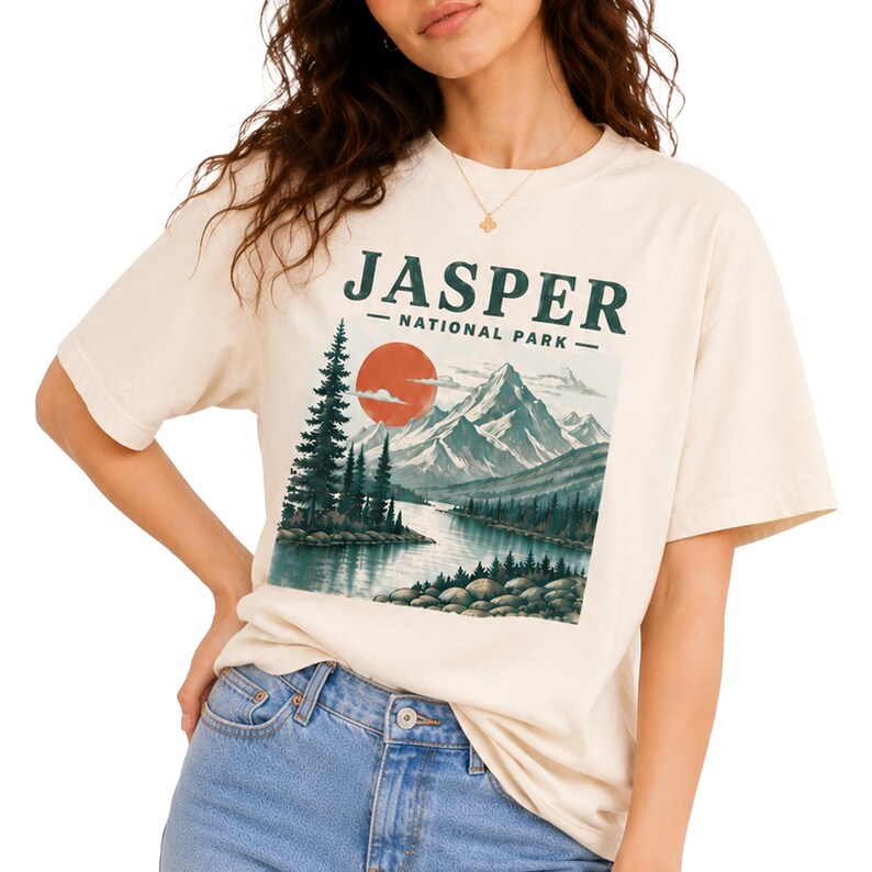 Pode incluir: Camiseta creme com uma ilustra&ccedil;&atilde;o paisag&iacute;stica do Parque Nacional Jasper. O design inclui montanhas, um rio, um sol poente e &aacute;rvores perenes. O texto "JASPER NATIONAL PARK" est&aacute; impresso acima do gr&aacute;fico.