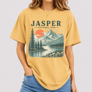 Pode incluir: Camiseta amarelo mostarda com uma ilustra&ccedil;&atilde;o c&ecirc;nica do Parque Nacional Jasper. O design inclui uma cordilheira, um lago, pinheiros e um sol poente. O texto "JASPER NATIONAL PARK" &eacute; impresso acima da paisagem.