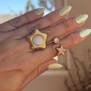 May include: A hand displaying three gold rings. One ring is a large star shape with a white center stone. Another is a small star with a pink stone, and the third is a small round ring with a clear stone.
