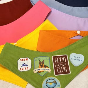May include: A collection of triangular pet bandanas in various solid colors including green, orange, yellow, pink, maroon, light blue, and navy. The green bandana features patches with text reading Snow Patrol, Fun Police, Good Dogs Club, Dad's Sidekick, and I'd Rather Be At The Lake.