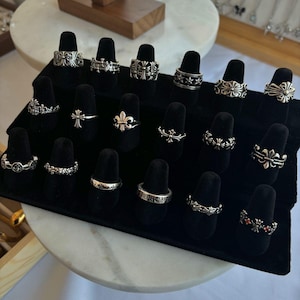 May include: A collection of twenty silver-toned rings displayed on a black velvet tiered stand. The rings feature various intricate designs, including crosses, fleur-de-lis motifs, and floral patterns, with one ring accented by small red stones.
