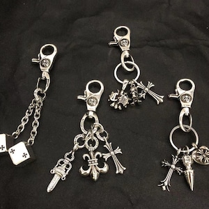 May include: Four silver-toned metal keychains featuring lobster claw clasps. One has two dice charms on chains, another has a dagger, fleur-de-lis, and cross charms, while the others feature various cross, skull, and heart-shaped metal charms.