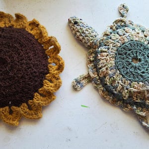 Puede incluir: Dos artículos de ganchillo hechos a mano: un girasol y una tortuga. El girasol tiene un centro marrón oscuro y pétalos amarillos. La tortuga es en tonos verdes, azules y beige. Ambos están sobre una superficie blanca.