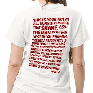 May include: White t-shirt with a crew neck and short sleeves. The back of the shirt features red text that reads "THIS IS YOUR NOT AT ALL HUMBLE REMINDER THAT SHANE, AKA, THE MAN. IS THE BADDEST BITCH IN THE MLH."