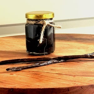 May include: A small glass jar with a gold lid, filled with dark liquid, sits on a wooden surface. Two dark brown vanilla beans are next to the jar. The jar is tied with twine.