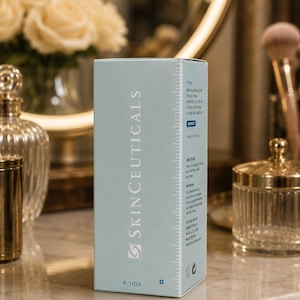 May include: A light blue box of SkinCeuticals P-Tiox peptide serum. The box has white text and a measurement scale on the side. The background includes a mirror, flowers, and cosmetic accessories.