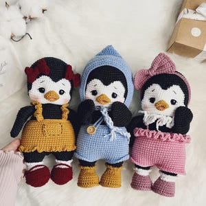Può includere: Tre bambole pinguino all'uncinetto fatte a mano con corpi neri e bianchi e becchi arancioni. Il pinguino a sinistra indossa una salopette giallo senape e scarpe rosse. Il pinguino centrale indossa un vestito azzurro con cappuccio e quello a destra rosa.