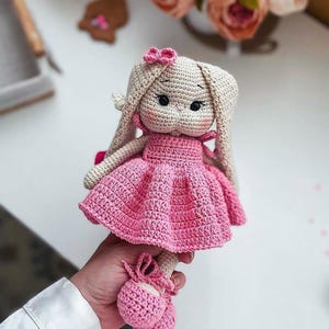 Può includere: Una bambola fatta all'uncinetto a mano con testa di coniglio beige, orecchie lunghe, occhi a perla neri e guance rosa. La bambola indossa un vestito rosa vivace con gonna svasata e scarpe all'uncinetto abbinate con nastri e un piccolo fiocco.