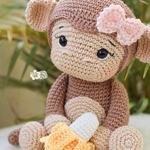 May include: A crocheted monkey doll with brown and beige yarn, featuring large black eyes with long lashes, a light pink bow on its head, and a small yellow and white crocheted banana held in its lap. The doll has a round head and limbs.
