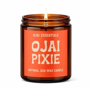 May include: A lit candle in a brown glass jar with a bright orange label. The label features the text Ojai Essentials, Ojai Pixie, and Natural Soy Wax Candle in a cream-colored font. The candle has a single wick burning with a small flame.