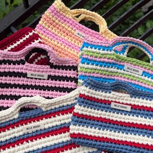 May include: A collection of five rectangular crochet handbags featuring horizontal stripes in various colour combinations including pink, yellow, blue, green, red, white, and black. Each bag has a white label with the text MILLIE BROOKE.
