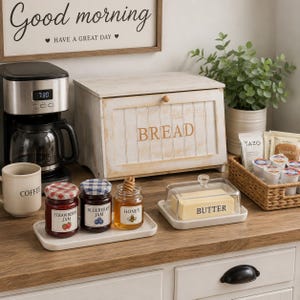 May include: A rustic white wooden bread box with the word BREAD printed on the front, a black coffee maker, a white mug labeled COFFEE, a white tray with strawberry jam, blueberry jam, and honey, and a glass butter dish labeled BUTTER.