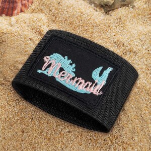 May include: Black wristband with a rectangular patch featuring a light blue mermaid tail and the word "Mermaid" in pink script. The wristband is made of a textured, woven material, and is set against a sandy background.