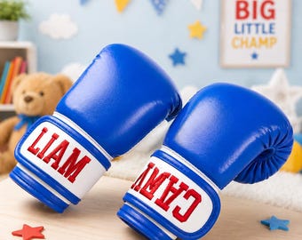 Op de afbeelding: Twee blauwe bokshandschoenen met witte polsbanden. De namen "LIAM" en "CAM" zijn in het rood op de handschoenen geborduurd. De handschoenen liggen op een licht oppervlak, met stervormige decoraties.
