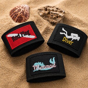 May include: Three black wristbands with embroidered patches. One reads "Red Flag" in red and white, another shows a diver silhouette with "Diver" in yellow, and the third says "Mermaid" in blue and pink. The wristbands are on a sandy surface.