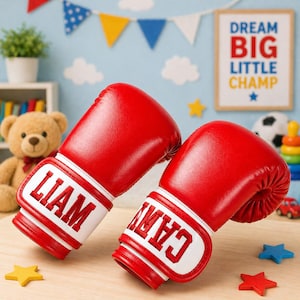 Peut inclure: Deux gants de boxe rouges avec des poignets blancs, l'un brodé du nom "LIAM" et l'autre "CAIN". Ils sont posés sur une surface marron clair. Un ours en peluche et une affiche "DREAM BIG LITTLE CHAMP" sont visibles en arrière-plan.