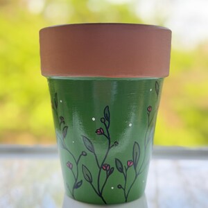 May include: A hand-painted green ceramic flower pot with a brown rim. The pot is decorated with black vines, pink flowers, and white dots. The pot is designed for indoor or outdoor use.