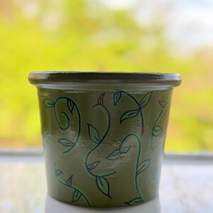 May include: A small, olive green ceramic plant pot with a dark green rim. The pot is decorated with a repeating pattern of stylized green vines and leaves. The pot is sitting on a white surface.