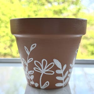 Pode incluir: Um vaso de flores de terracota castanho com desenhos florais brancos. O vaso apresenta flores e folhas brancas pintadas à mão no exterior. O vaso foi concebido para plantar flores e outras plantas.