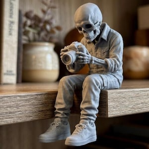 Może przedstawiać: Szara figurka szkieletu w koszuli, dżinsach i trampkach, trzymającego aparat fotograficzny. Szkielet siedzi na drewnianej półce, z książkami i elementami dekoracyjnymi w tle.