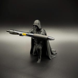 May include: A black figurine of Darth Vader holding a black and yellow Pilot G-2 0.7 pen. The figurine is standing on a black surface. The pen has the text "PILOT G-2 0.7" printed on it.