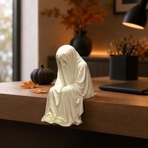 May include: A white ghost figurine with a draped sheet appearance, featuring a sad facial expression with downturned eyes and a small mouth. The figure is sculpted in a seated position with its legs hanging over the edge of a flat surface.