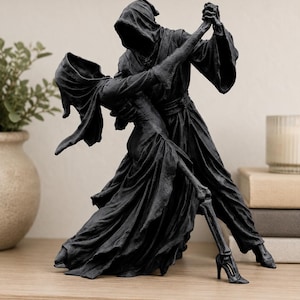 May include: A matte black sculpture depicting two hooded figures in a dance pose. One figure wears a long, flowing robe, while the other wears a dress and high-heeled shoes. The figures have skeletal hands and feet, with one leg clearly showing bone structure.