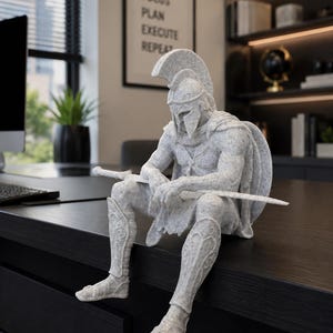 May include: A light gray speckled stone-effect figurine of a seated Spartan warrior wearing a crested helmet and armor, holding a long sword across the lap. The warrior has a shield on the back and detailed greaves on the legs, sitting in a resting pose.