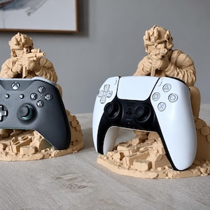 May include: Two beige monochromatic soldier figurines acting as gaming controller stands. One soldier holds a black Xbox controller, while the other holds a white PlayStation 5 controller. Both figures are sculpted in a kneeling position on rubble.