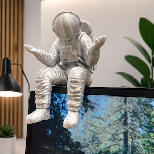 May include: A silver-colored astronaut figurine with arms outstretched, seated on the edge of a laptop. The astronaut is wearing a detailed spacesuit with a helmet and backpack. The laptop screen displays a forest scene.