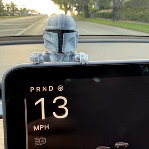 Op de afbeelding: Een grijze 3D-geprinte Mandalorian helmfiguur zit op het dashboard van een auto. Het display van de auto toont de versnellingskeuze, de snelheid van 21 km/u en andere rij-informatie. De achtergrond toont een weg en bomen.