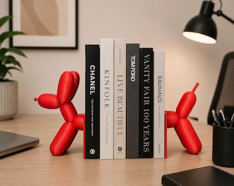 May include: A pair of bright red balloon dog bookends flanking a row of five books. The books feature titles including Chanel Collections and Creations, Kinfolk, Live Beautiful, Tom Ford, Vanity Fair 100 Years, and Bauhaus Magdalena Droste.