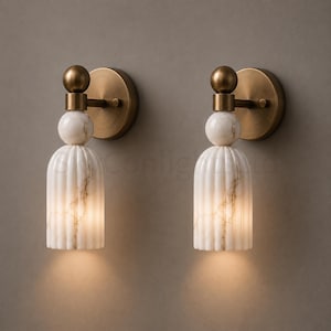 Puede incluir: Dos apliques de pared con acabado de latón y detalles de mármol blanco. Cada aplique presenta una placa trasera redonda de latón, una esfera de mármol y una pantalla de mármol acanalada en forma de campana. Los apliques están iluminados.