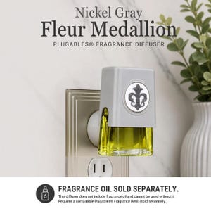 May include: A Nickel Gray Fleur Medallion Plugables fragrance diffuser plugged into a wall outlet. The device features a light gray rectangular housing with a circular emblem displaying a dark gray fleur-de-lis design and a clear glass reservoir.