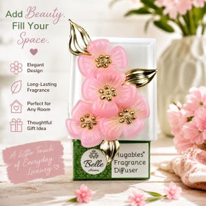 May include: A white plug-in fragrance diffuser featuring three translucent pink flowers with gold centers and gold leaves. The base is green with white text reading Belle Aroma Plugables Fragrance Diffuser. Text on the left includes Add Beauty. Fill Your Space. Elegant Design, Long-Lasting Fragrance, Perfect for Any Room, Thoughtful Gift Idea, and A Little Touch of Everyday Luxury.