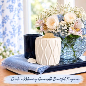 May include: A cream-colored ceramic wax warmer with a vertical wave pattern and a wax melt on top, positioned next to a dark blue glass candle holder. The text "Create a Welcoming Home with Beautiful Fragrance" is displayed at the bottom.