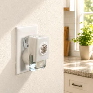 May include: A white electric wall plug-in fragrance warmer featuring a rectangular body with a circular gold-toned filigree medallion on the front. The device is plugged into a standard wall outlet and includes a clear glass refill bottle.
