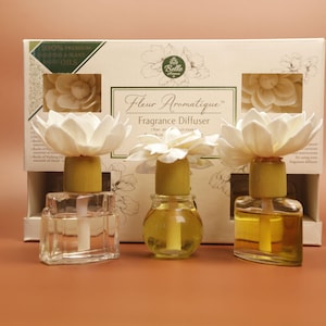 May include: Three glass fragrance diffusers with white flower-shaped toppers and wooden collars, positioned in front of a white box labeled Fleur Aromatique Fragrance Diffuser. The bottles contain clear and yellow liquids with visible internal wicks.